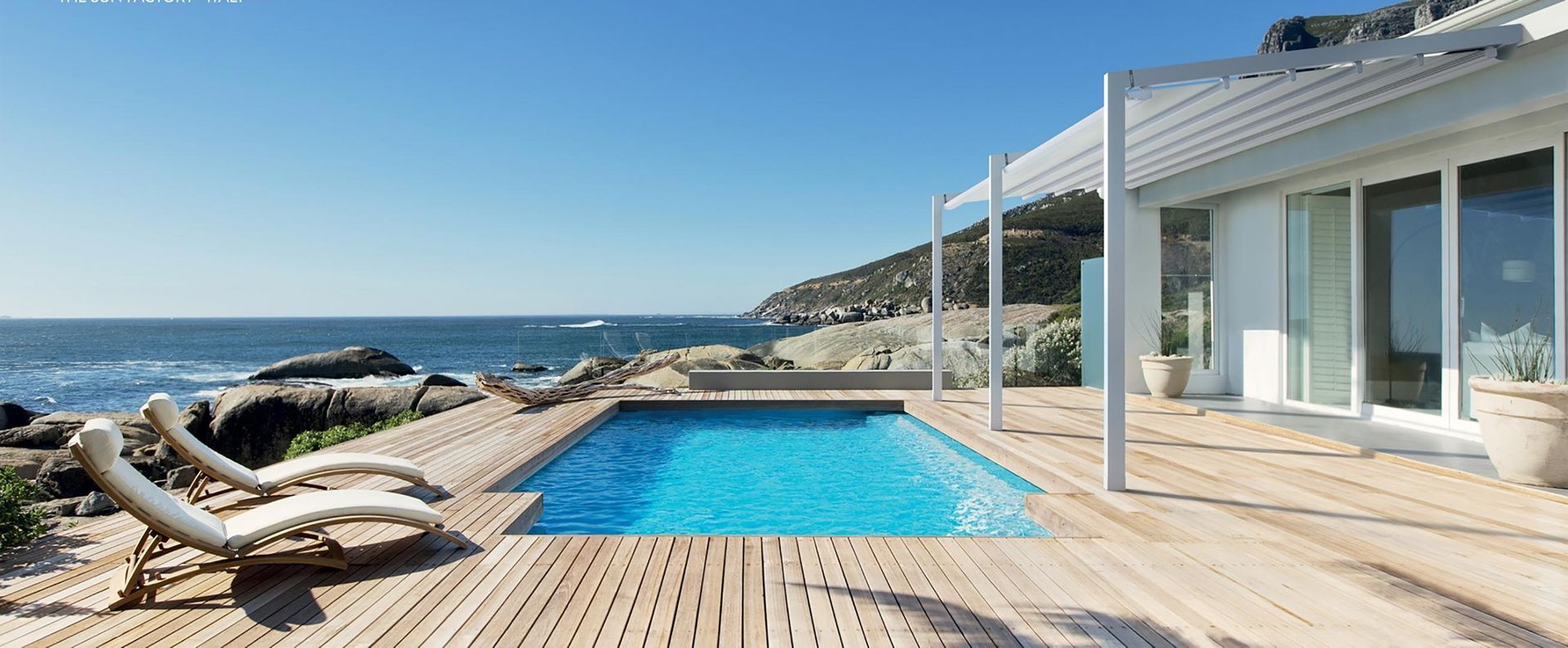 Patio fronte oceano con piscina blu, terrazza in legno, sedie a sdraio e moderna casa bianca con vista sul mare.