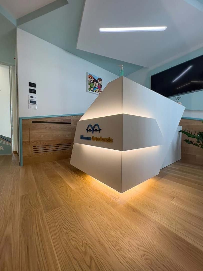 Moderna hall d'ingresso per uffici con bancone reception geometrico bianco e pavimento in legno, illuminata da una calda luce dal basso.