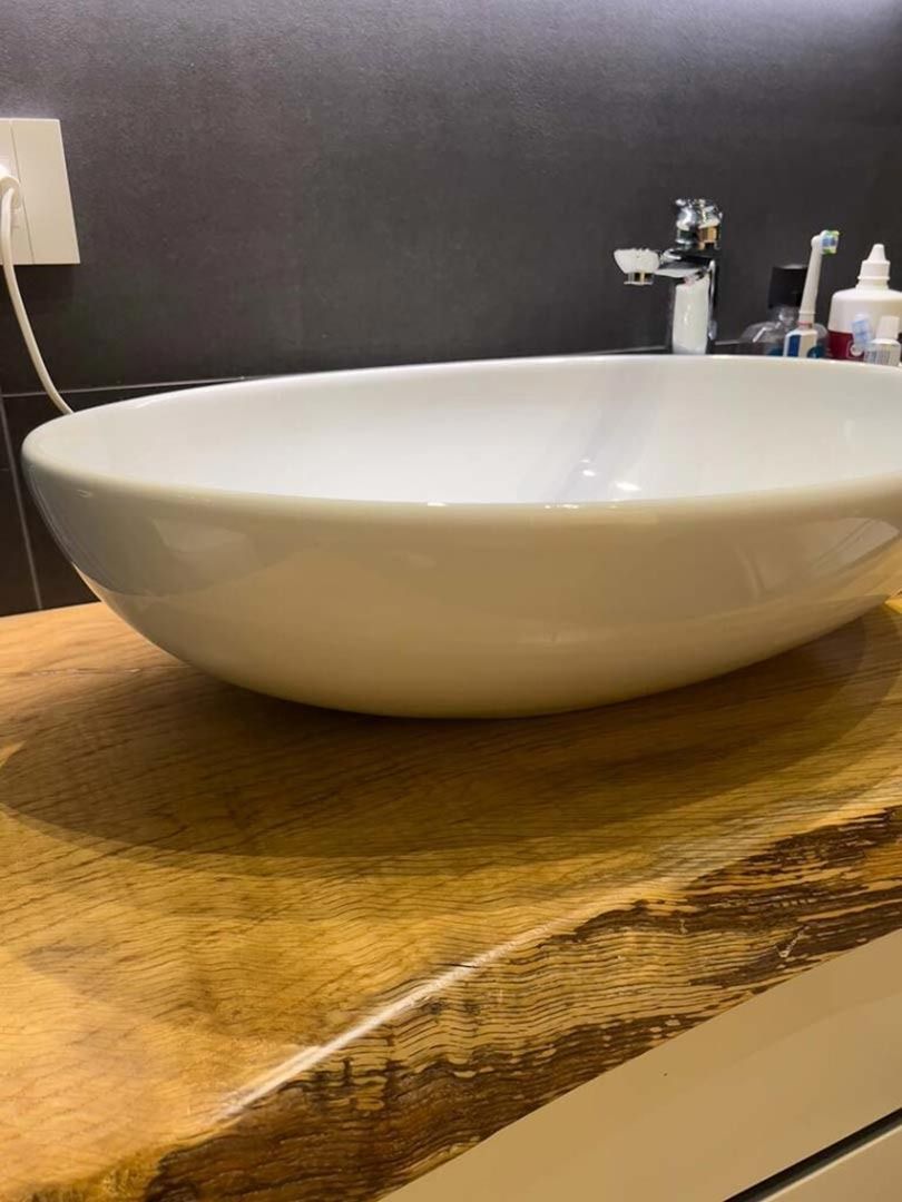 Lavabo ovale bianco su un piano di lavoro in legno con rubinetto da bagno e parete piastrellata di scuro alle sue spalle.