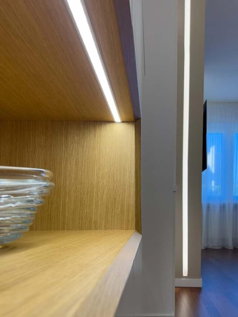 Mensola in legno con ciotola di vetro, illuminata da una sottile striscia LED; corridoio e tende sullo sfondo.