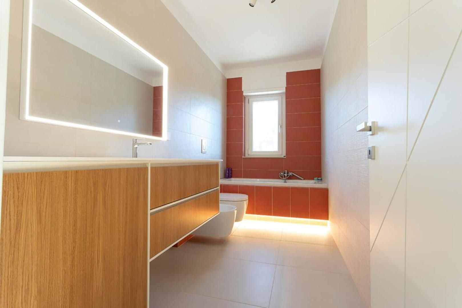Bagno moderno con mobile lavabo in legno, specchio, vasca da bagno e parete rivestita di piastrelle rosse accanto a una finestra.
