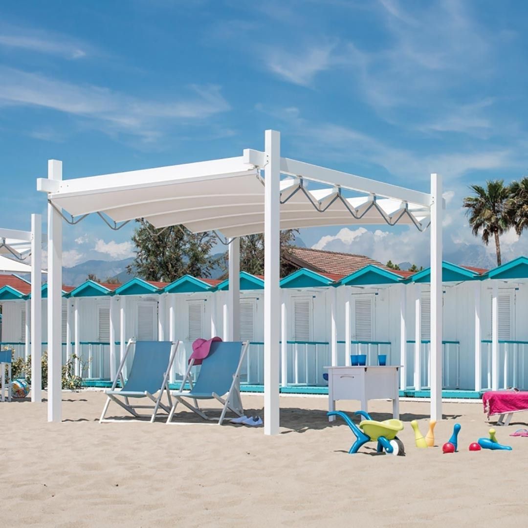 Un gazebo bianco su una spiaggia soleggiata con due sedie blu, giochi nella sabbia e ombrelloni turchesi dietro una recinzione.