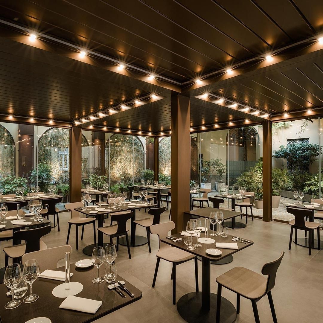Elegante sala da pranzo del ristorante con tavoli apparecchiati, luci soffuse dal soffitto e ampie finestre che si affacciano sul verde.