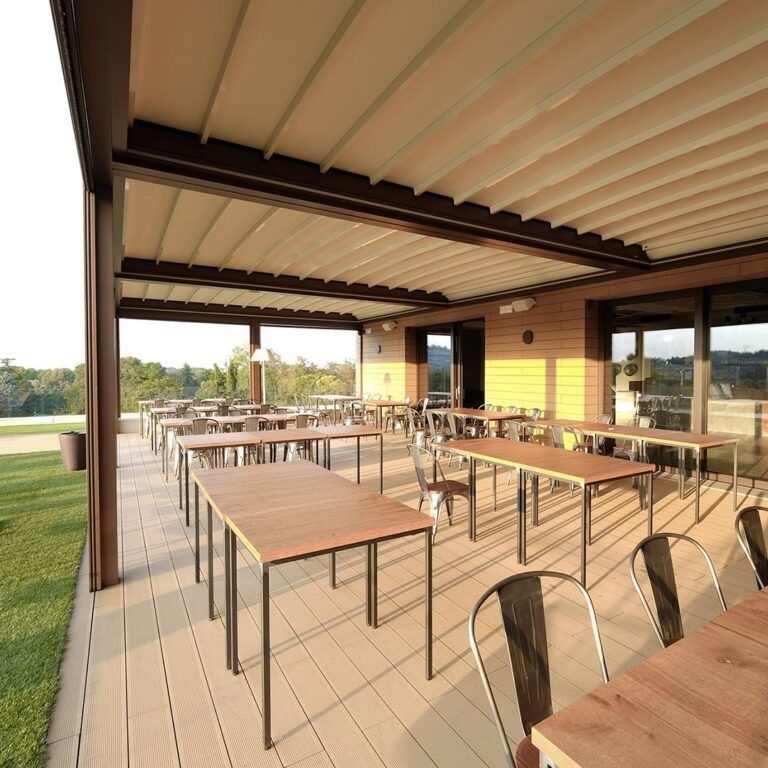 Zona pranzo esterna coperta con tavoli in legno, sedie in metallo e vista aperta sulla campagna al tramonto.