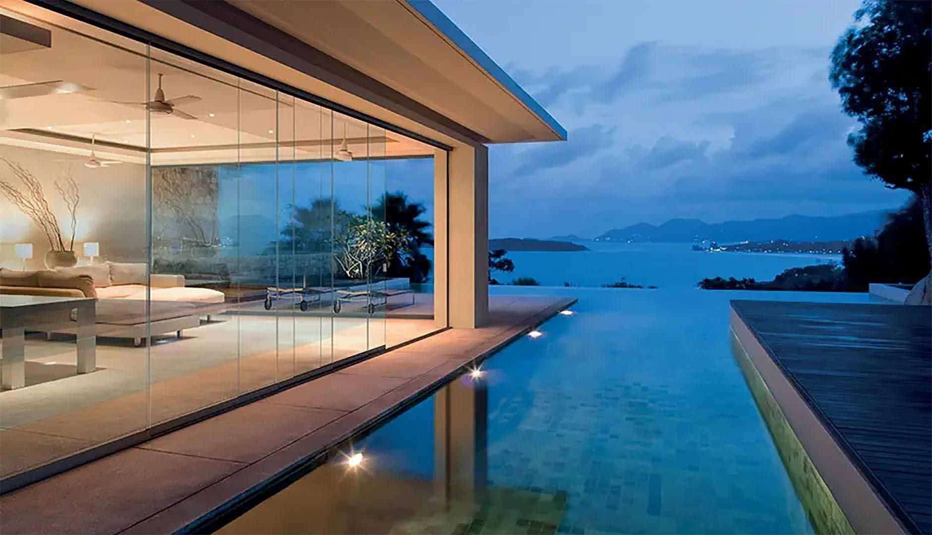 Villa moderna con pareti in vetro e piscina a sfioro affacciata su un oceano tranquillo al tramonto.