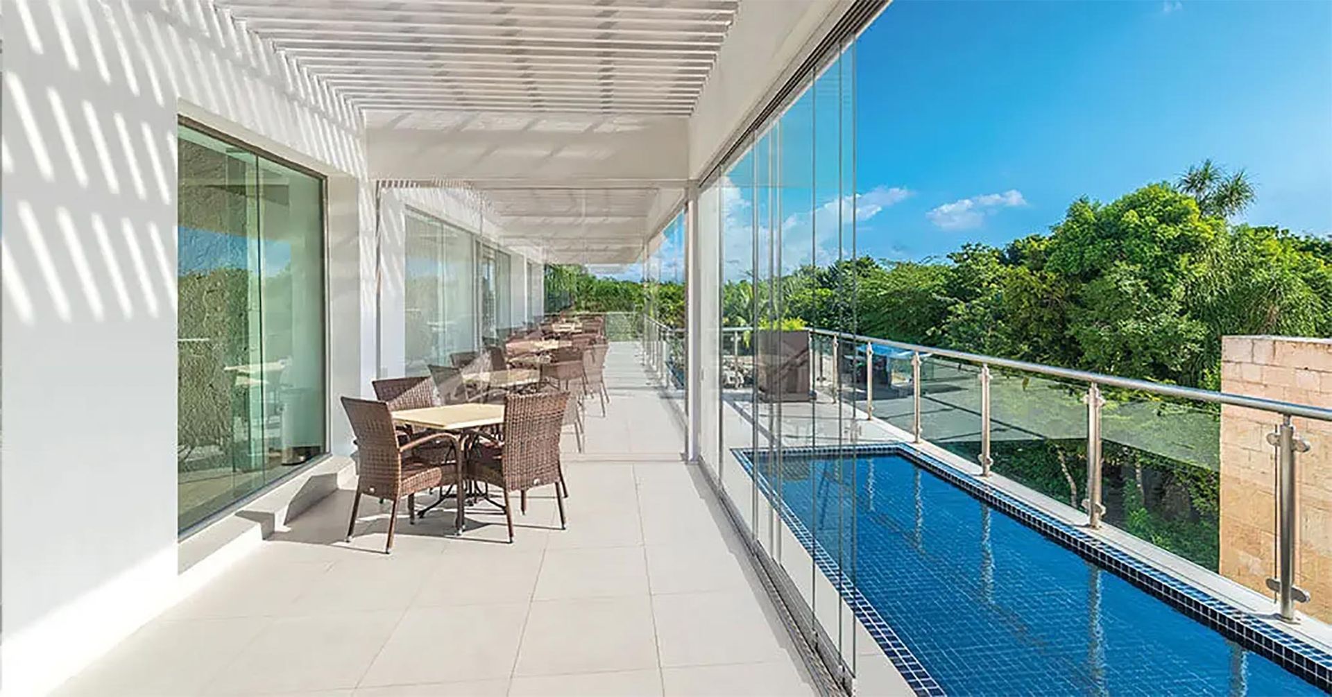Moderno balcone con set da pranzo all'aperto, ringhiera in vetro e piscina a sfioro blu con vista sul verde lussureggiante.