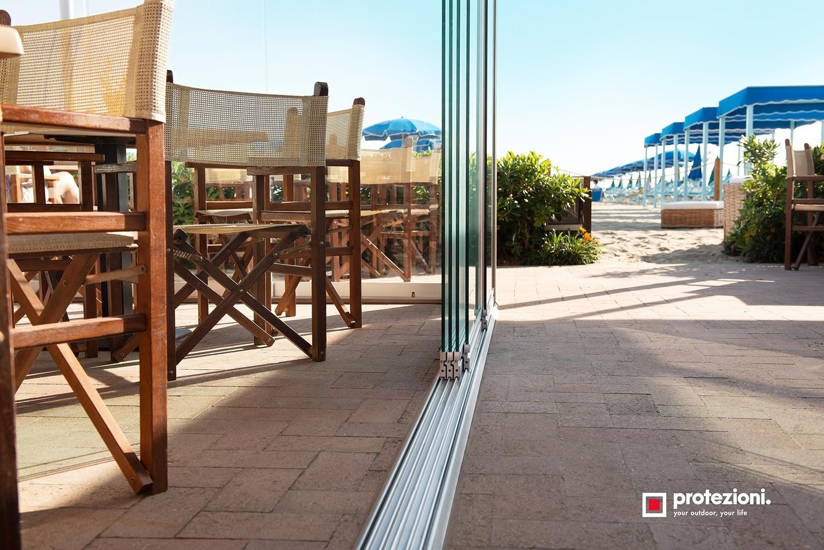 Patio esterno con sedie e tavoli in legno accanto a una porta a vetri, con vista su un vialetto soleggiato e tende da sole blu.