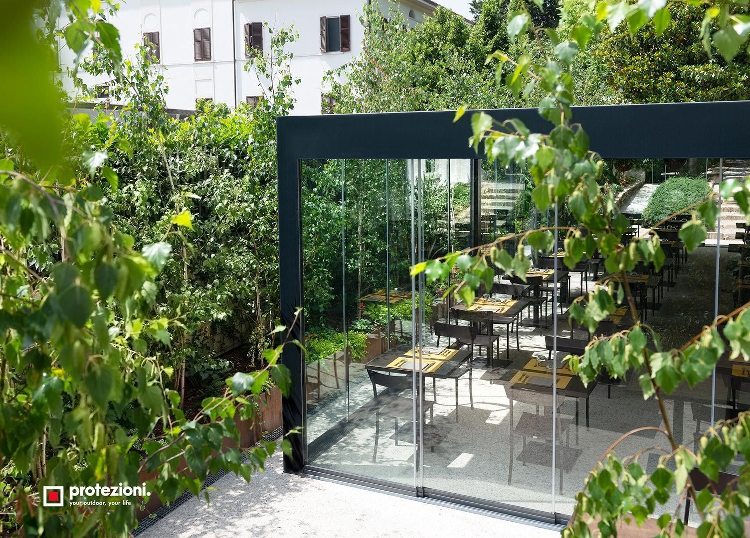 Caffè nel cortile dietro un cancello di metallo nero, con piante verdi e tavolini all'aperto baciati dal sole.