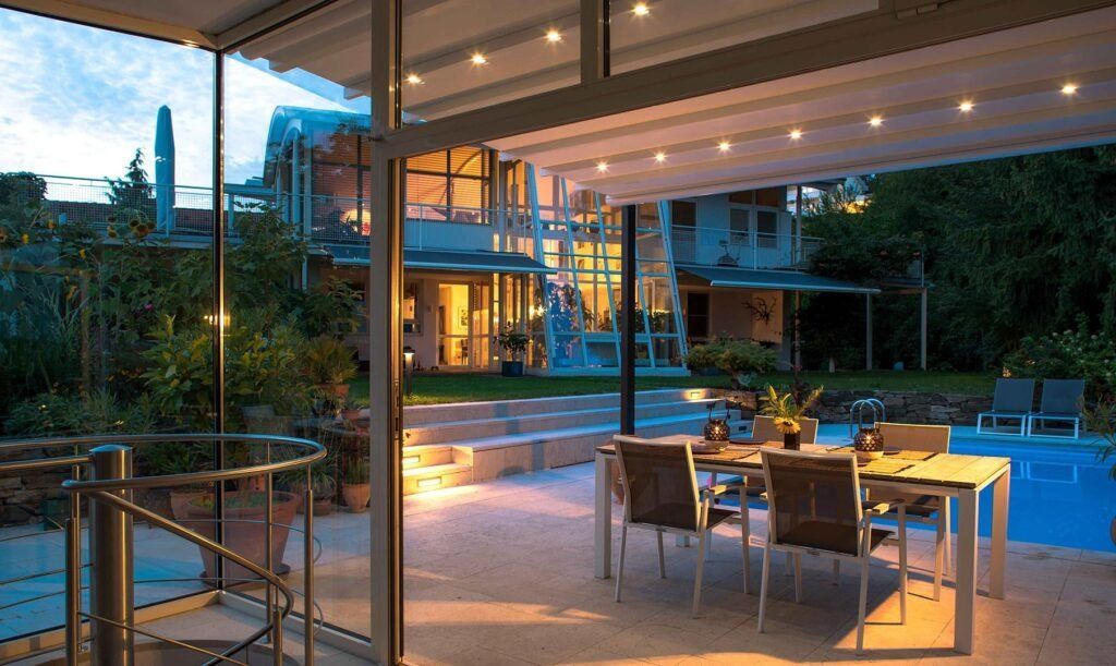Patio moderno con tavolo da pranzo e sedie a sdraio accanto a una piscina illuminata al crepuscolo.