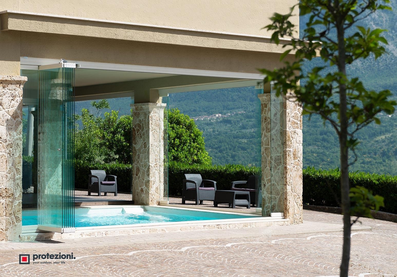 Patio moderno con piscina con pareti in vetro, colonne in pietra, lettini prendisole e vista sulle montagne.