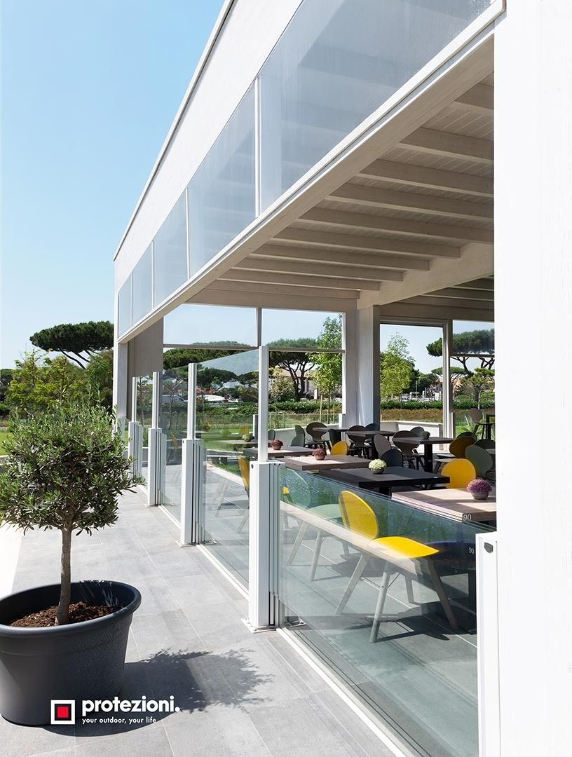 Terrazza ristorante moderna all'aperto con ringhiera in vetro, tettoia bianca e sedie gialle che si affaccia sul verde.