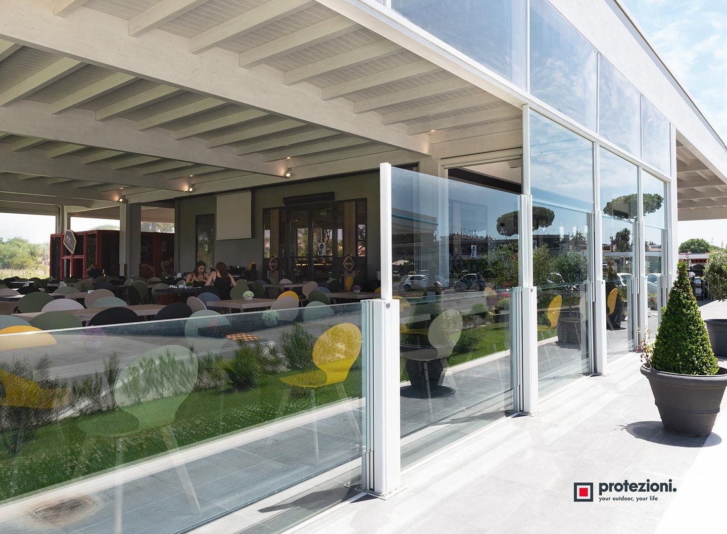 Ristorante moderno con patio dalle pareti in vetro, posti a sedere all'aperto e piante in vaso sotto una terrazza coperta.