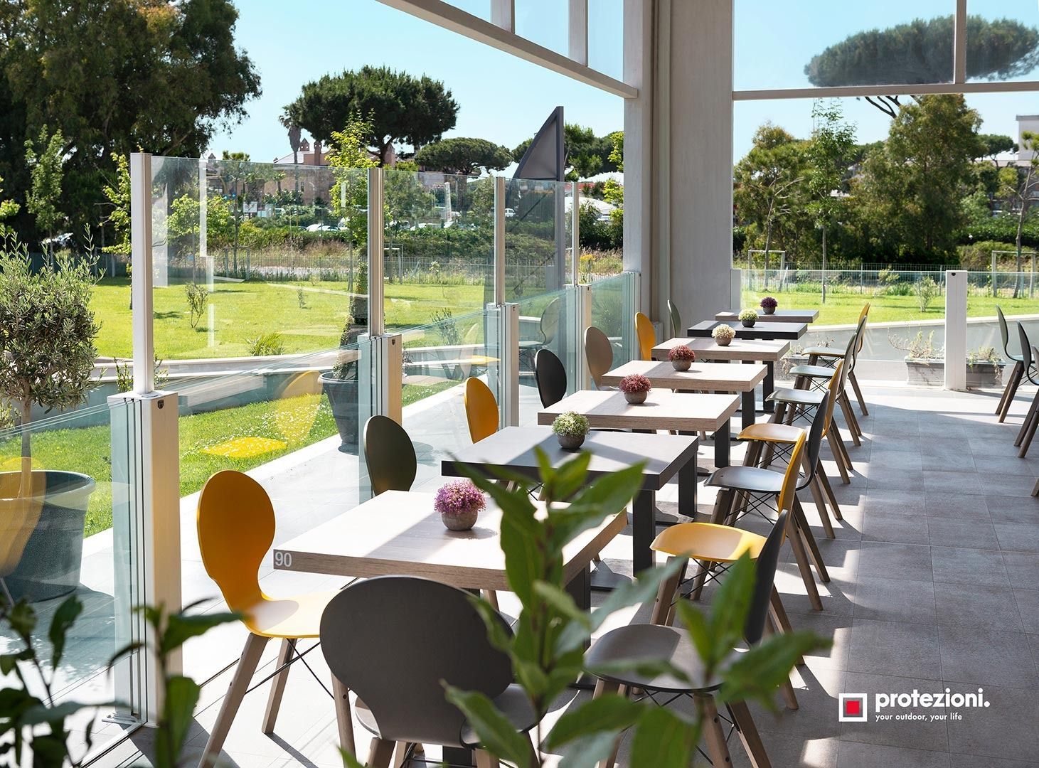 Luminoso caffè moderno con tavoli e sedie bianchi accanto a grandi finestre che si affacciano su un giardino verde.