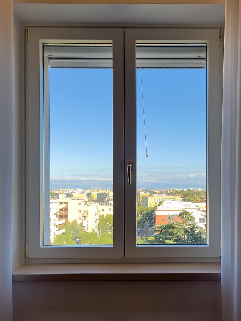 Finestra aperta con vista sulla città e cielo azzurro oltre il balcone.
