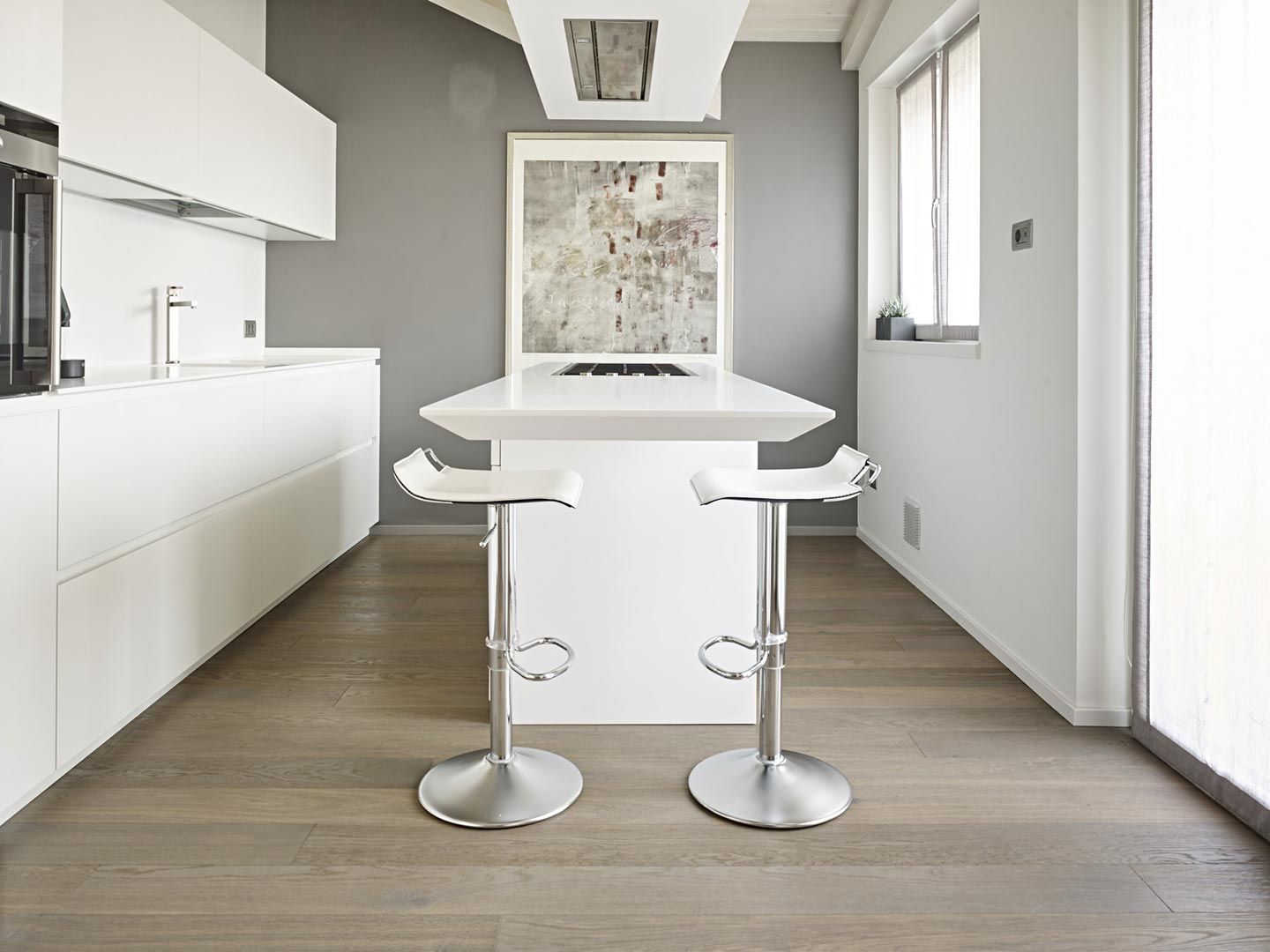 Cucina moderna bianca con isola centrale, due sgabelli e pavimento in legno chiaro.