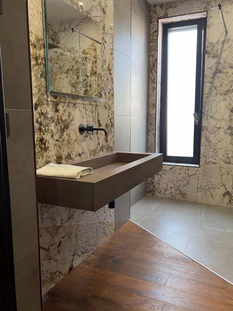 Bagno moderno con lavabo sospeso, finestra stretta, pareti effetto marmo e pavimento in legno.