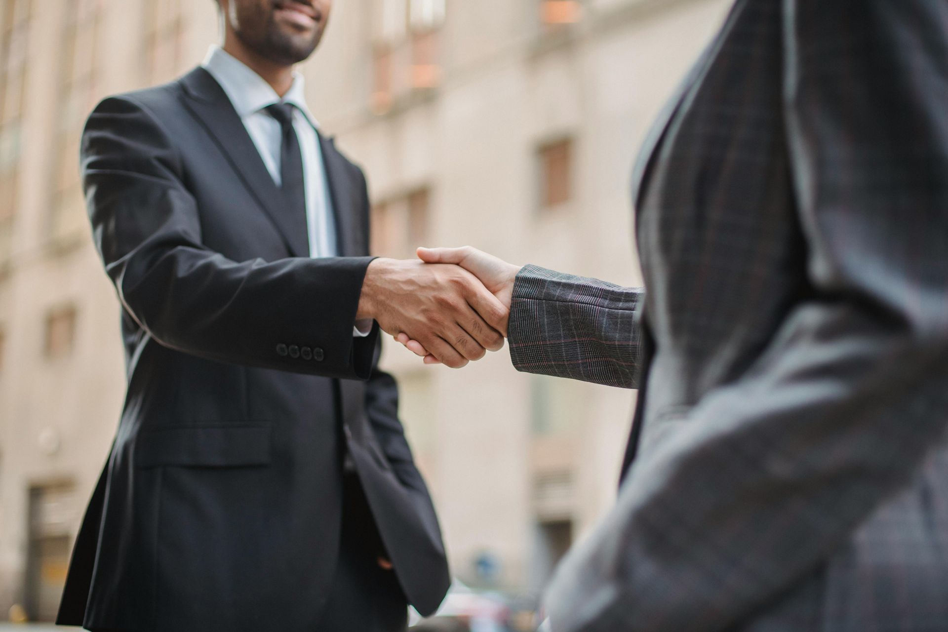 Two business professionals in suits shaking hands outdoors against a blurred architectural background.