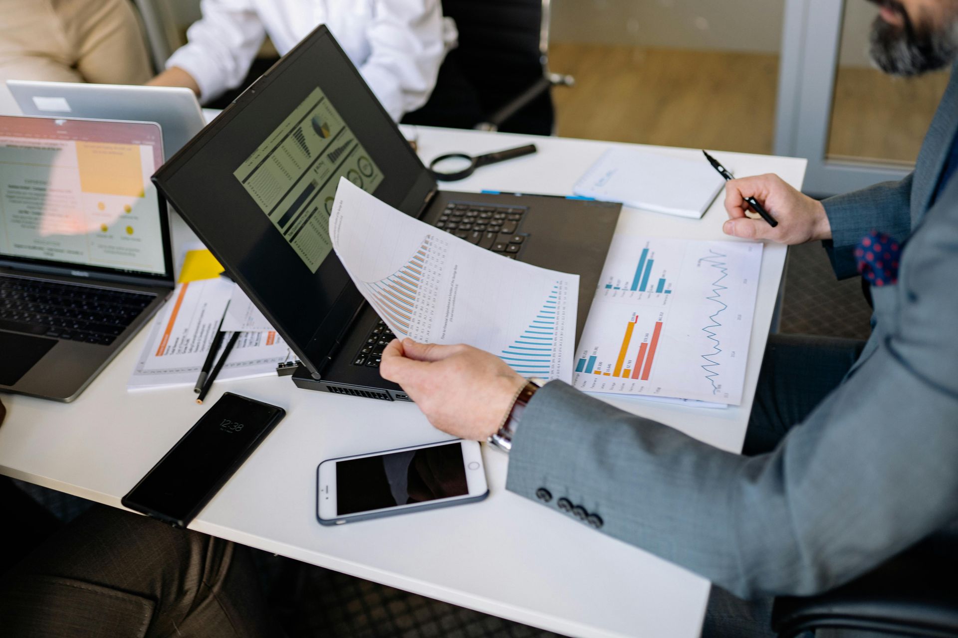 A person in a business suit reviewing financial charts and documents at a desk with multiple laptops and smartphones.