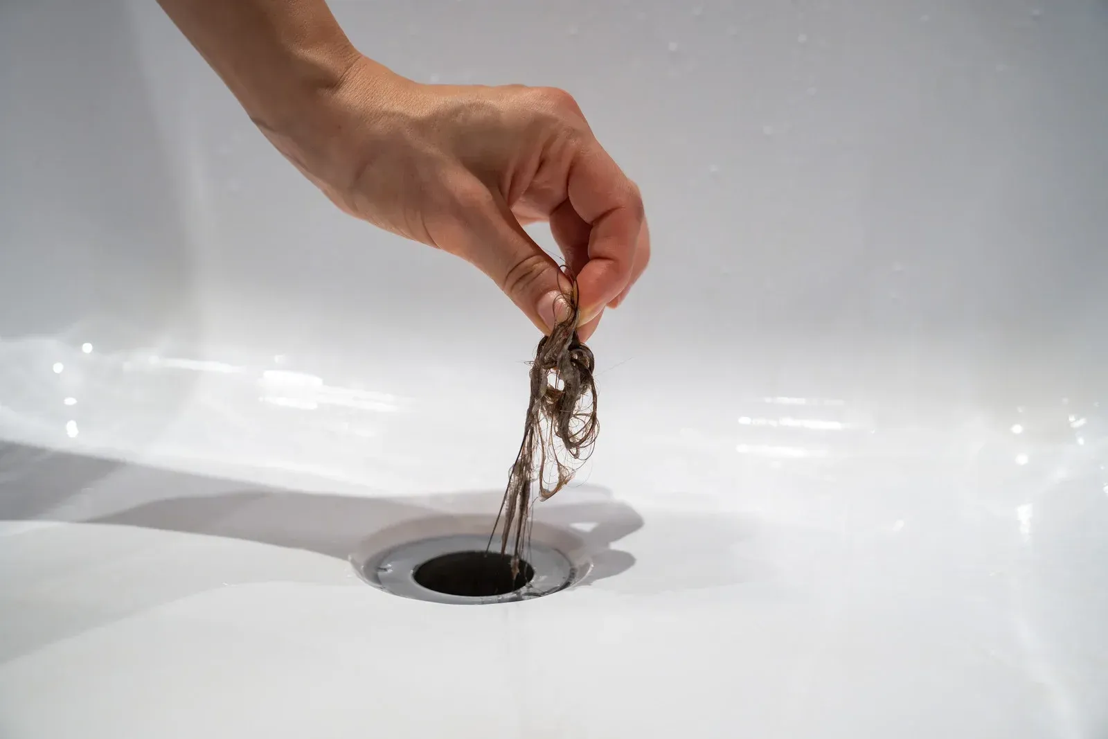 Hand holding a clump of hair over a drain in a white bathtub.