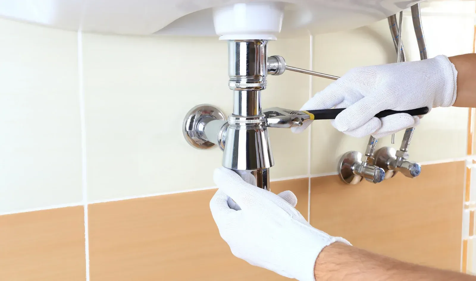 Plumber in white gloves using a wrench to work on a chrome sink drain in a bathroom.