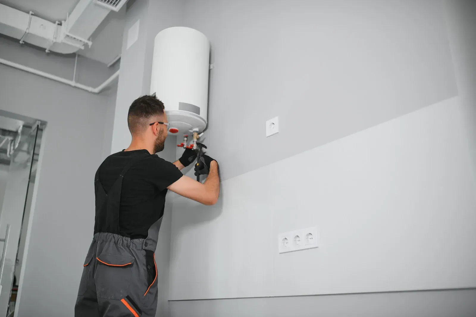 A technician wearing work coveralls uses a tool to install a white wall-mounted electric water heater.