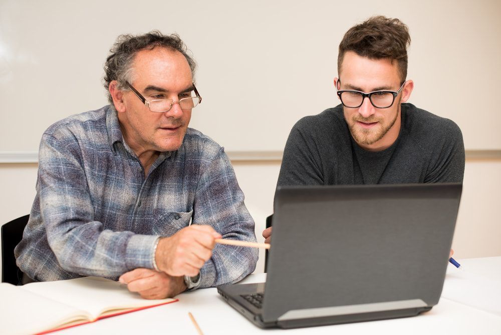 Two Men Are Sitting At A Table Looking At A Laptop Computer — Black Label I.T. in Garbutt, QLD