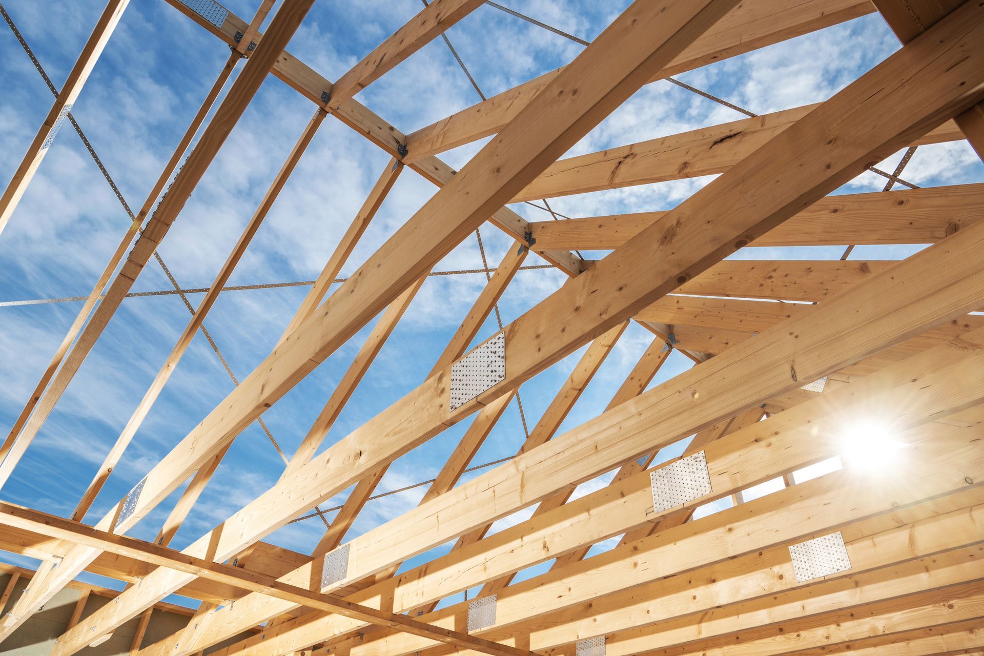 Wooden roof trusses under construction against a blue sky, with sunlight streaming through.