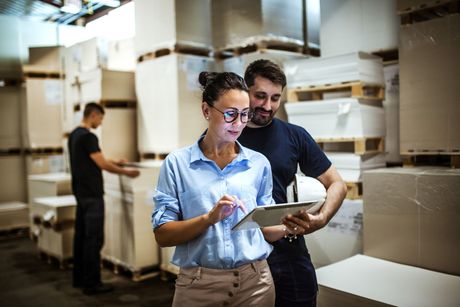 Two workers reviewing a tablet in a warehouse aisle among stacked boxes.