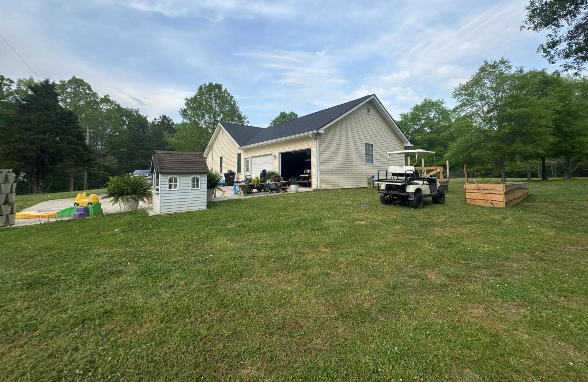 Beige house with open garage door on a grassy lawn. Includes a playhouse, golf cart, and wooden planter.
