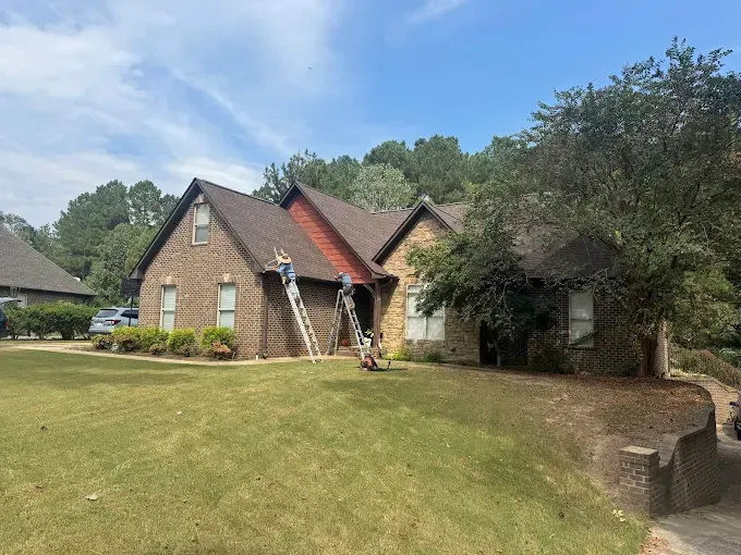 Two people on ladders are working on a brick house roof under a blue sky.