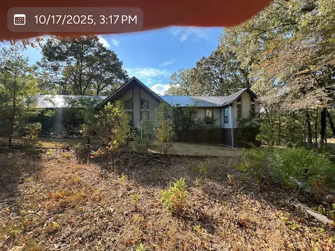 House with dark roof, surrounded by trees and brush, on a sunny day.