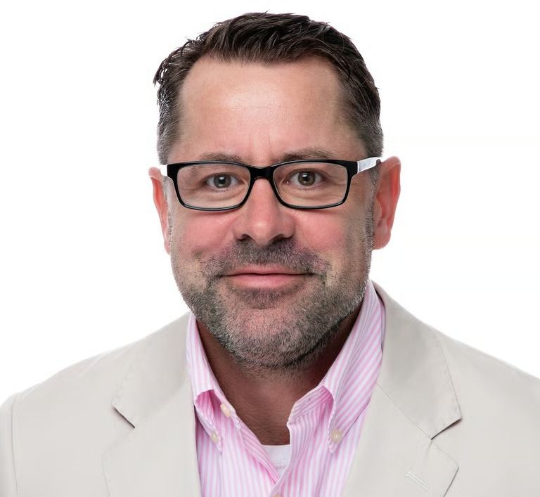 Man with glasses wearing a light jacket and pink striped shirt, smiling.