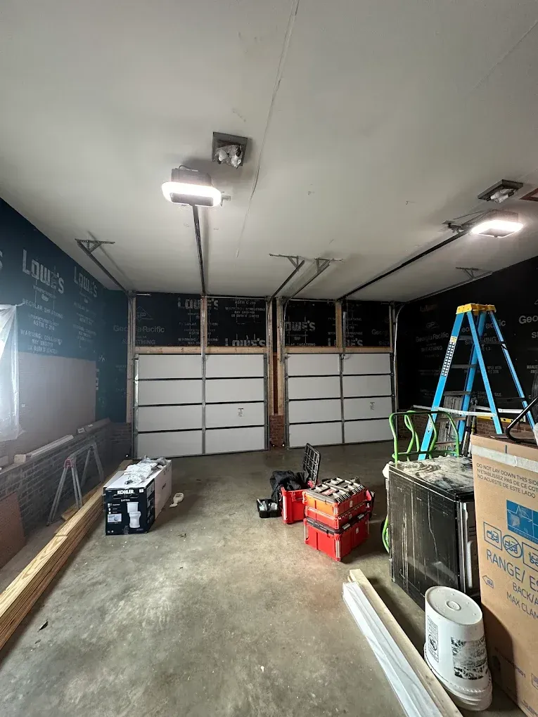 Unfinished garage with open garage door, ladder, tools, boxes, and construction materials on the floor.