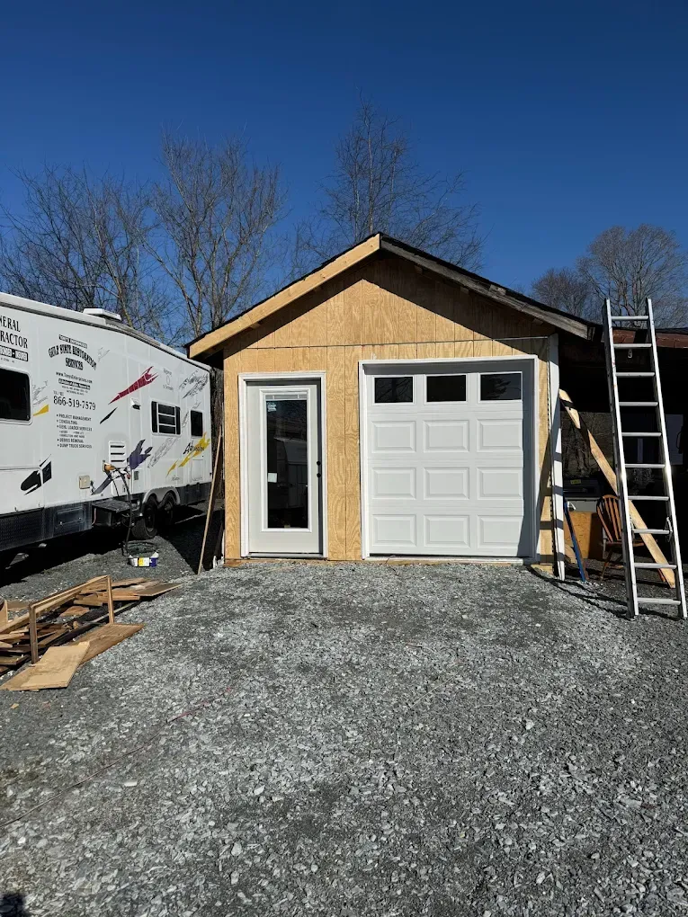 Small unfinished garage with a white door and window, gravel driveway, and ladder on the right.