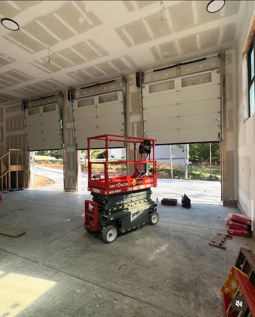 Red scissor lift inside an unfinished garage with open roll-up doors and construction materials.