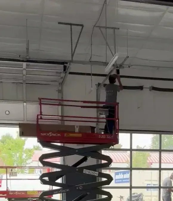 Worker standing on a red scissor lift, reaching up to adjust overhead ceiling fixtures in a warehouse.