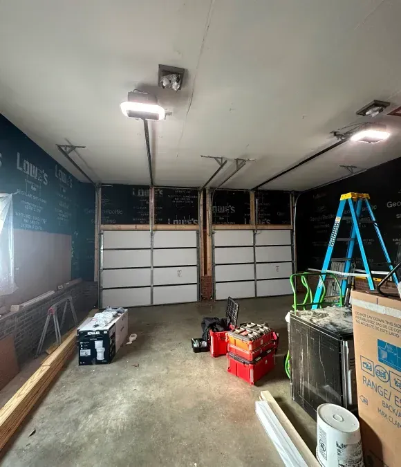 Cluttered garage interior with closed white doors, tools, ladder, and boxes on the floor