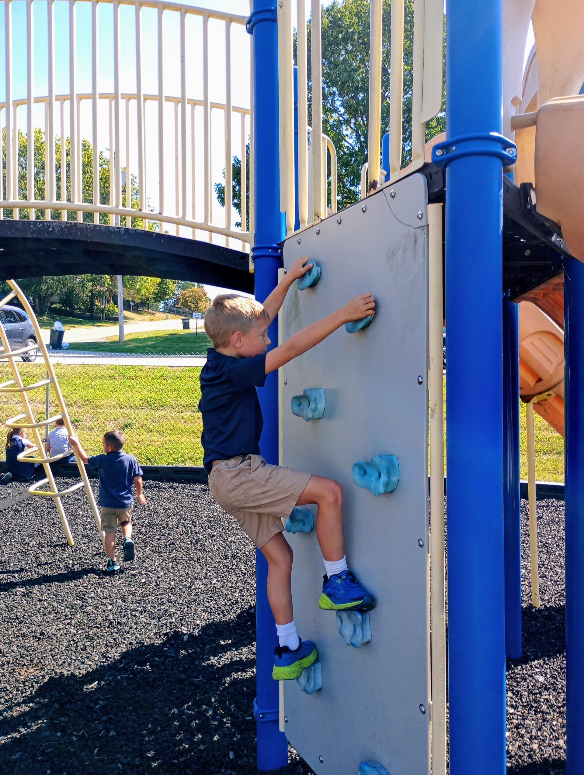 Child climbing a blue playground wall while another child climbs nearby on a sunny playground