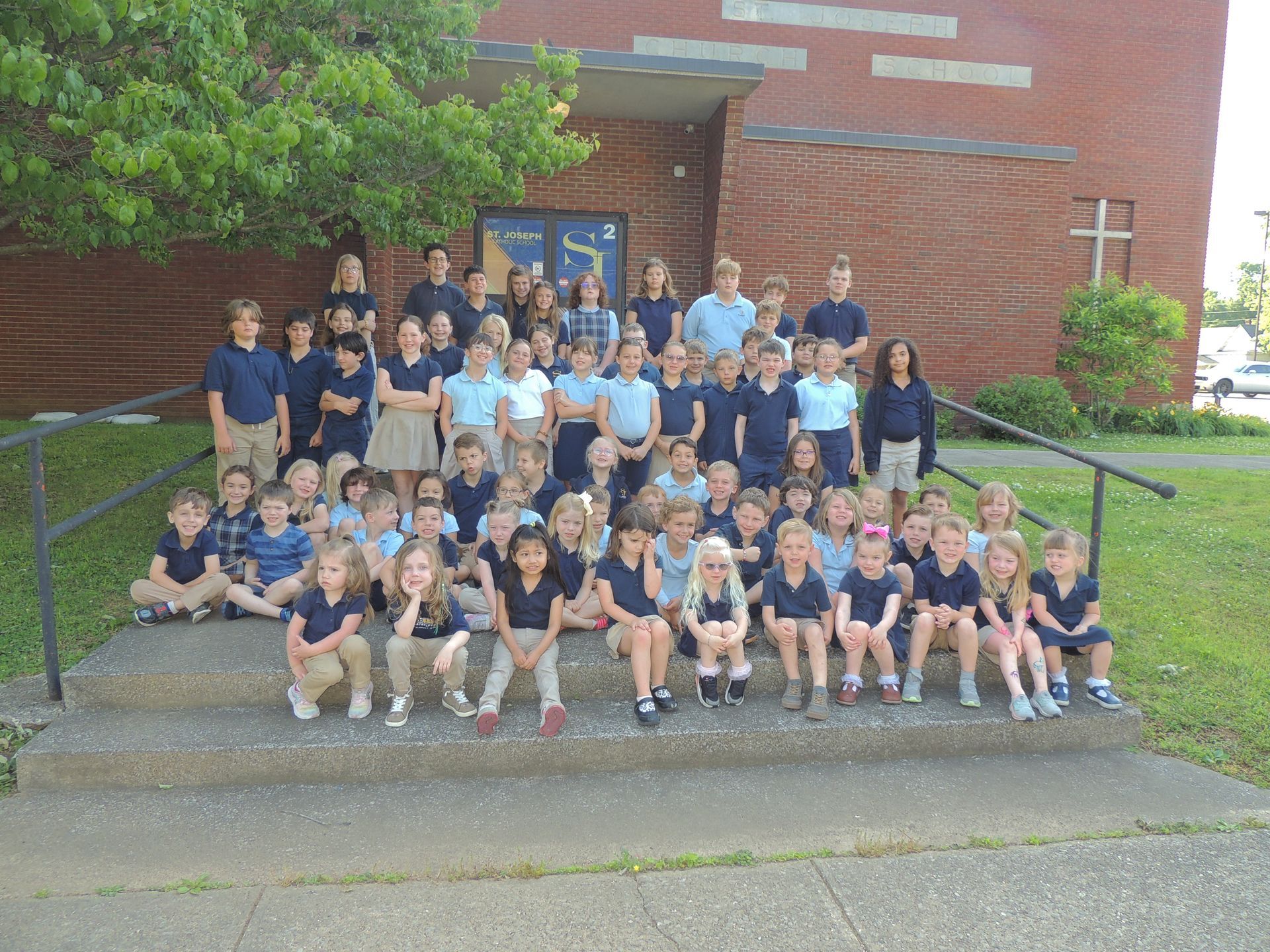 Large group posing on school steps outside a brick building with green trees nearby