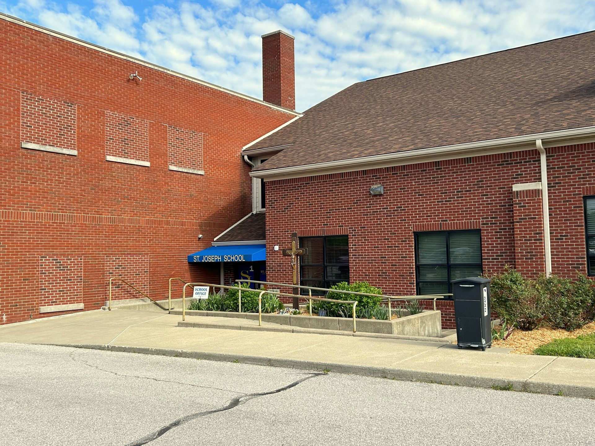 Brick building entrance with blue awning, windows, and paved driveway under a cloudy sky