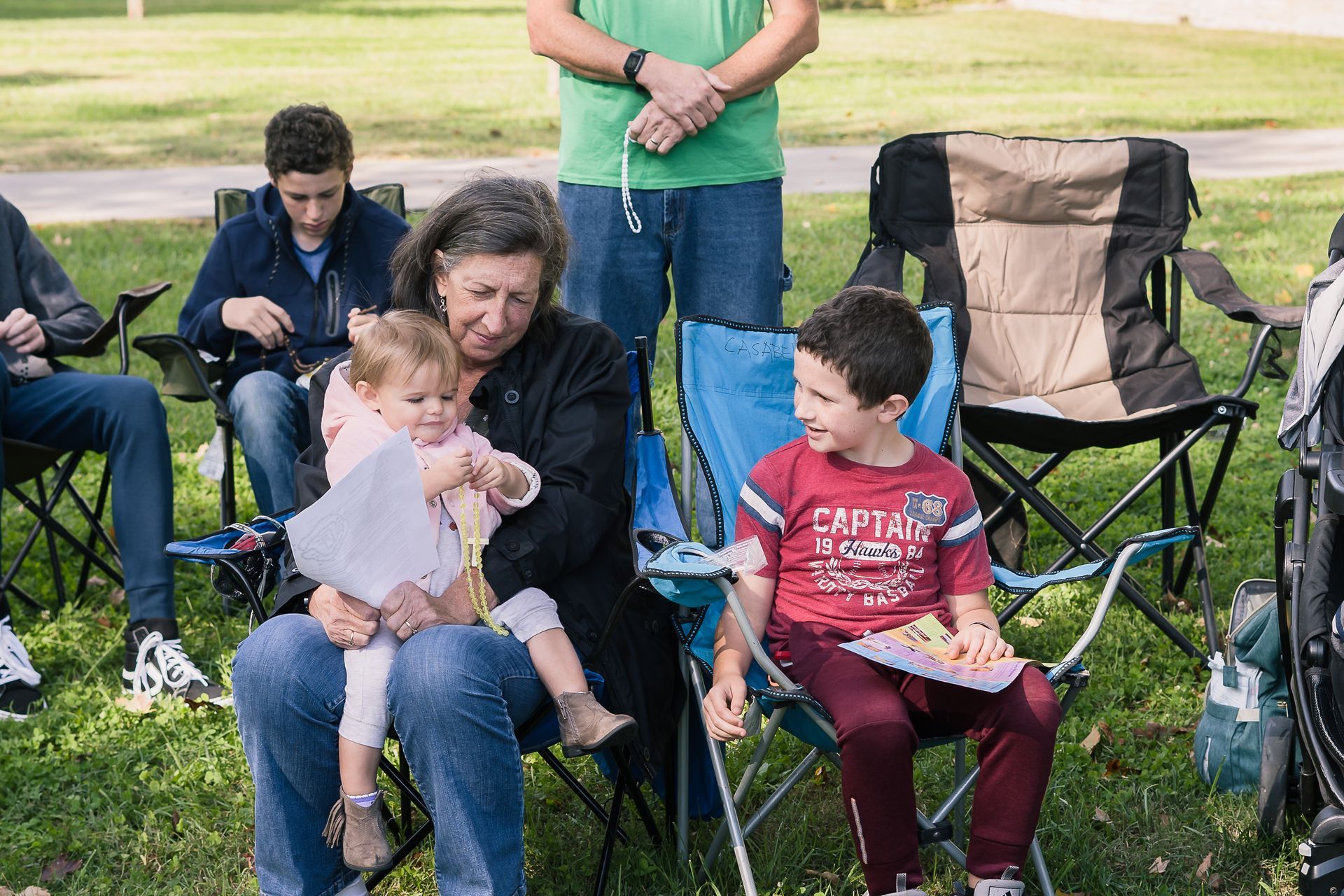 People seated in folding chairs at an outdoor park, one woman holding a child while others chat.
