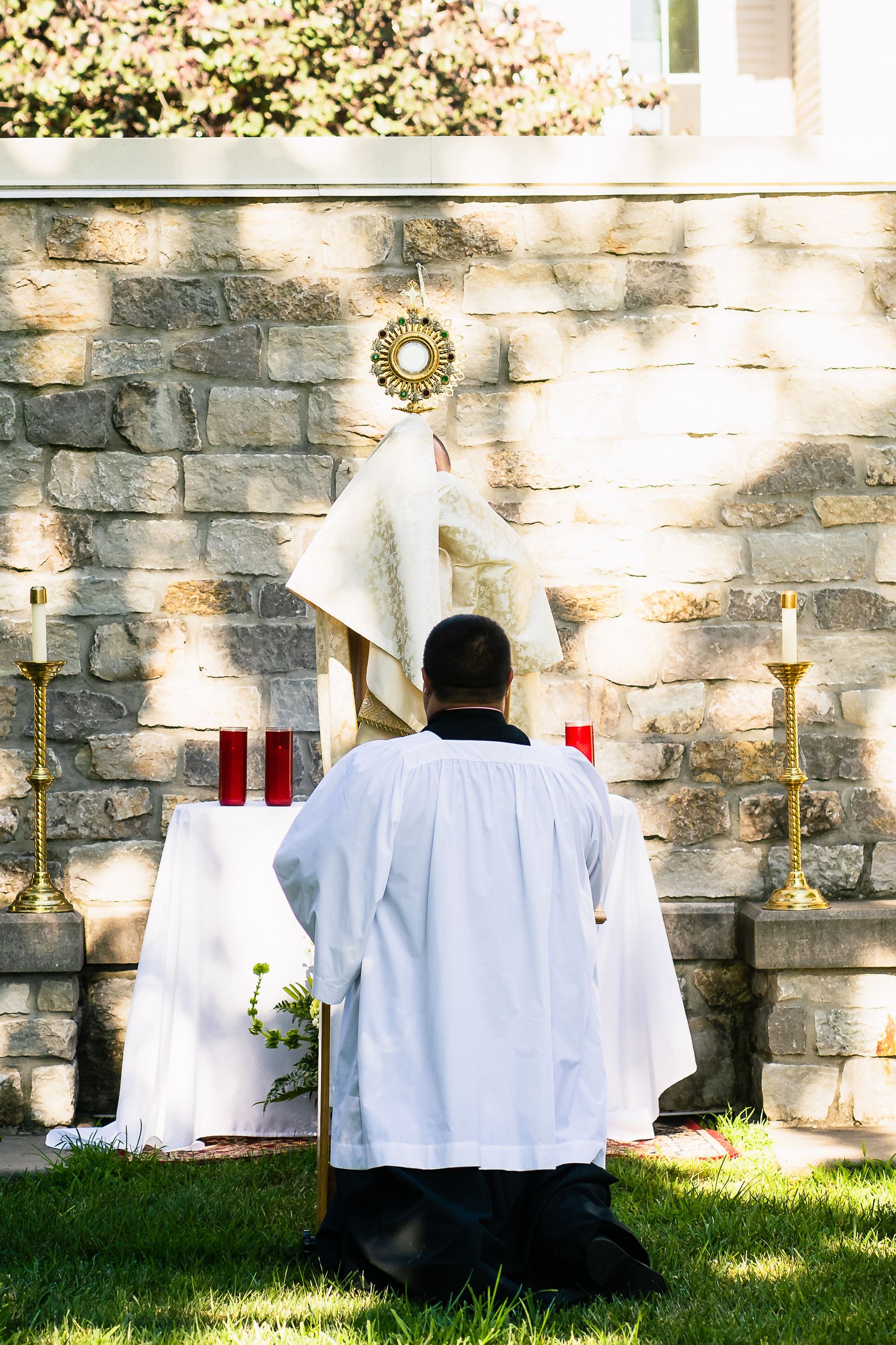 Person kneeling before an altar outdoors, with white cloths, candles, and a stone wall backdrop.