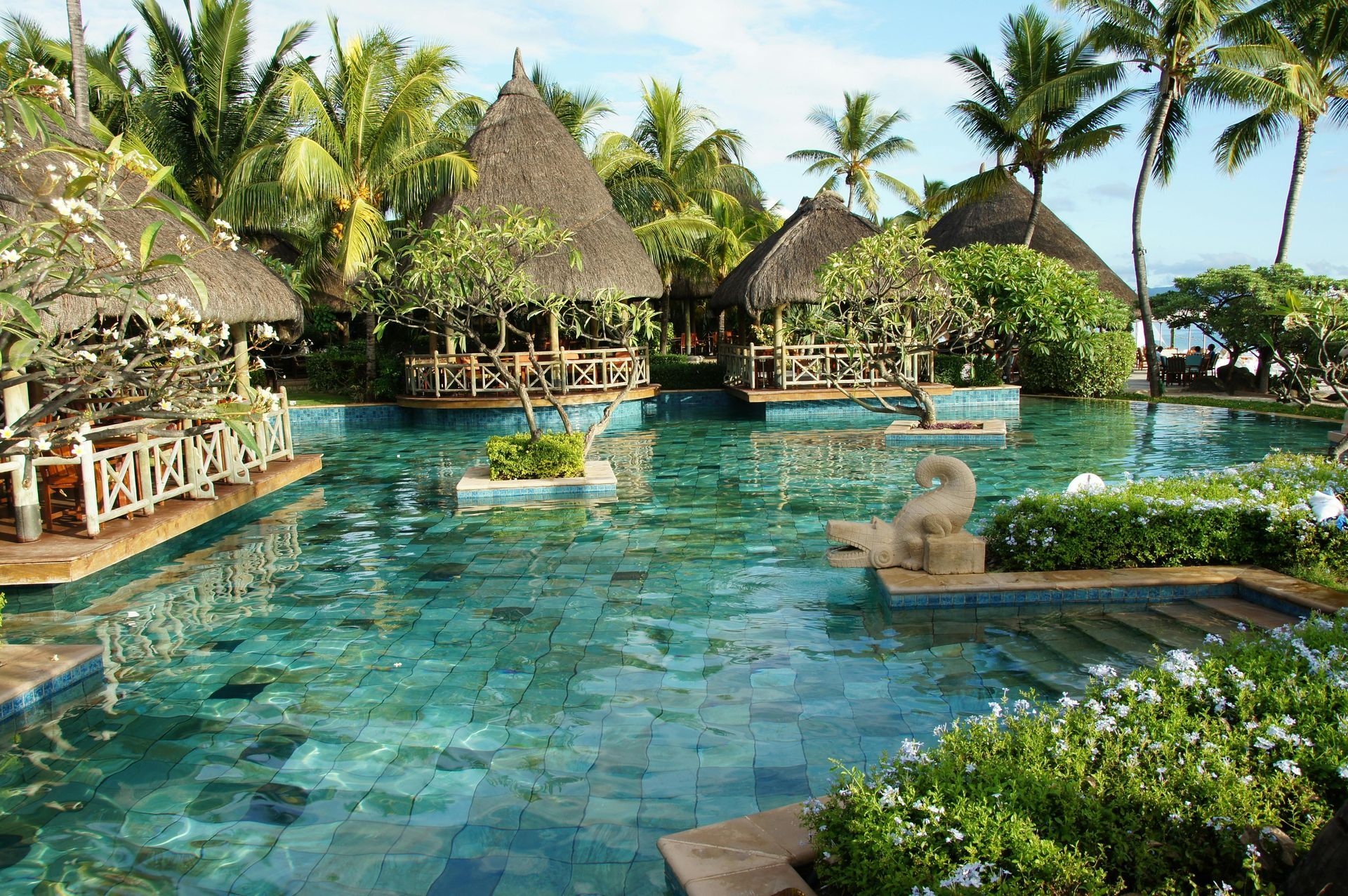 A tropical resort swimming pool with turquoise water, surrounded by lush green foliage and thatched-roof pavilions.