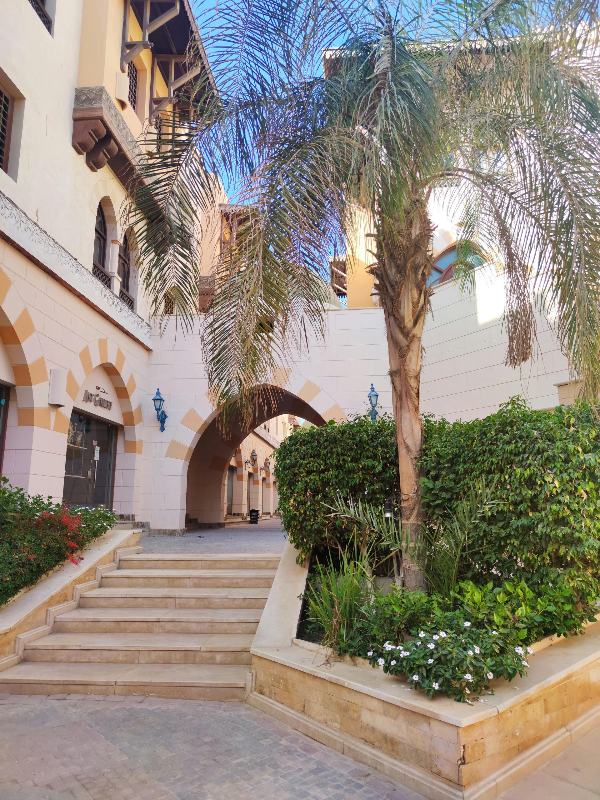 A staircase leads to an arched walkway between cream-colored buildings with Mediterranean architecture and palm trees.