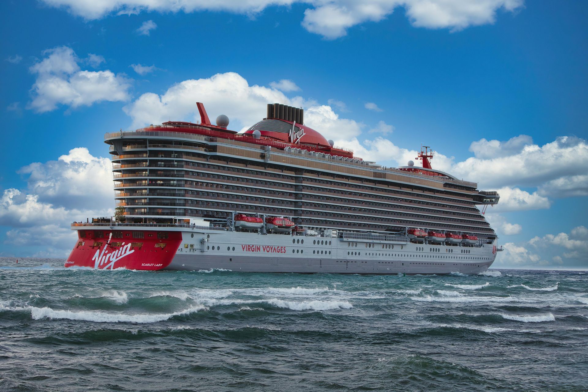 A large Virgin Voyages cruise ship with a red hull and red smokestack sailing on blue ocean waters under a cloudy sky.