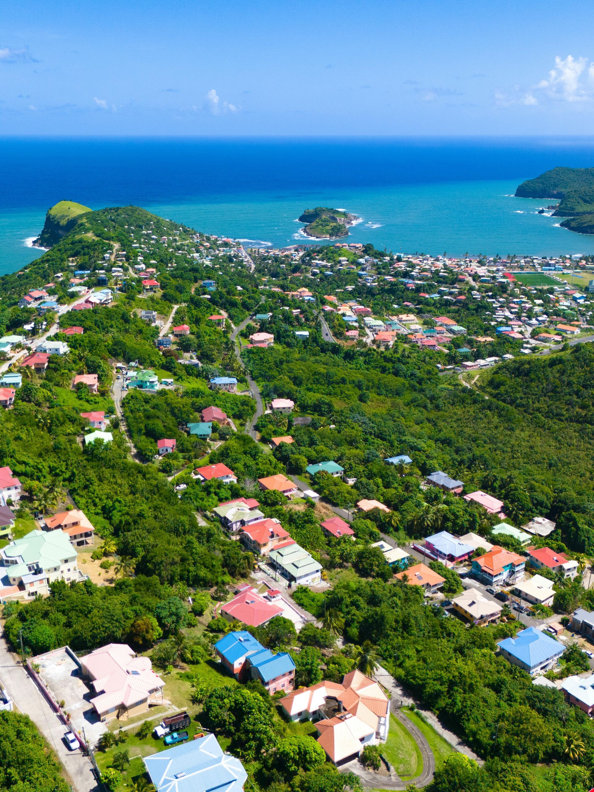Aerial view of houses nestled in a lush, green hillside sloping down toward a scenic tropical bay with blue water.
