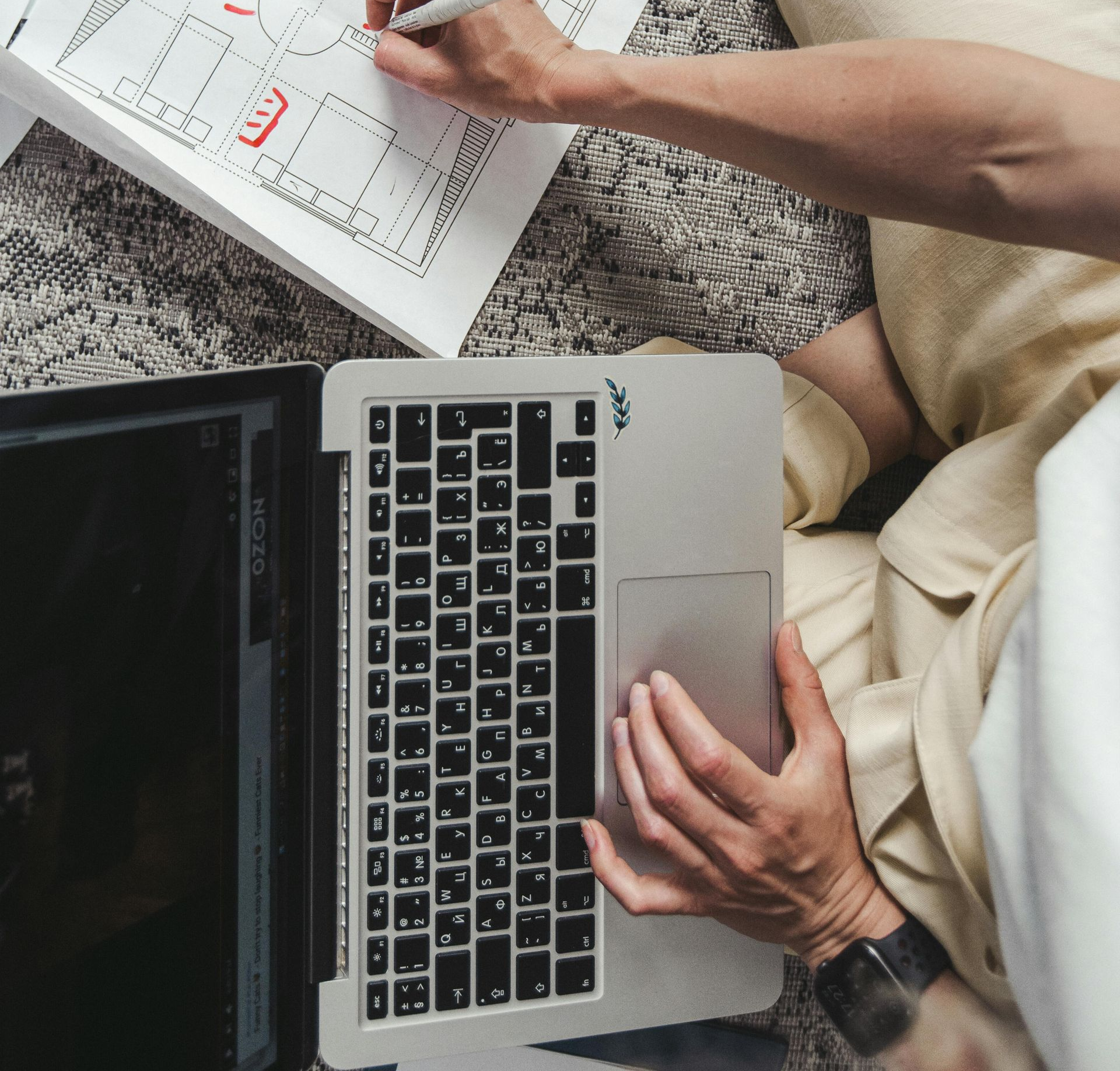 A person in light-colored clothing uses a laptop and marks a printed architectural floor plan with a pen.