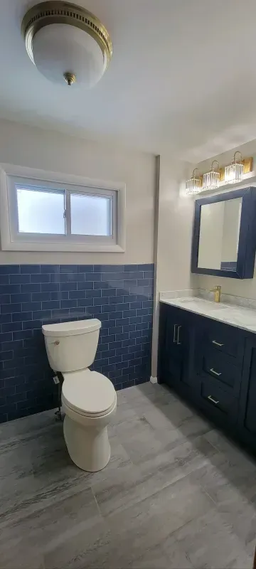 Small bathroom with white toilet, blue tile wainscoting, vanity, mirror, and frosted window.
