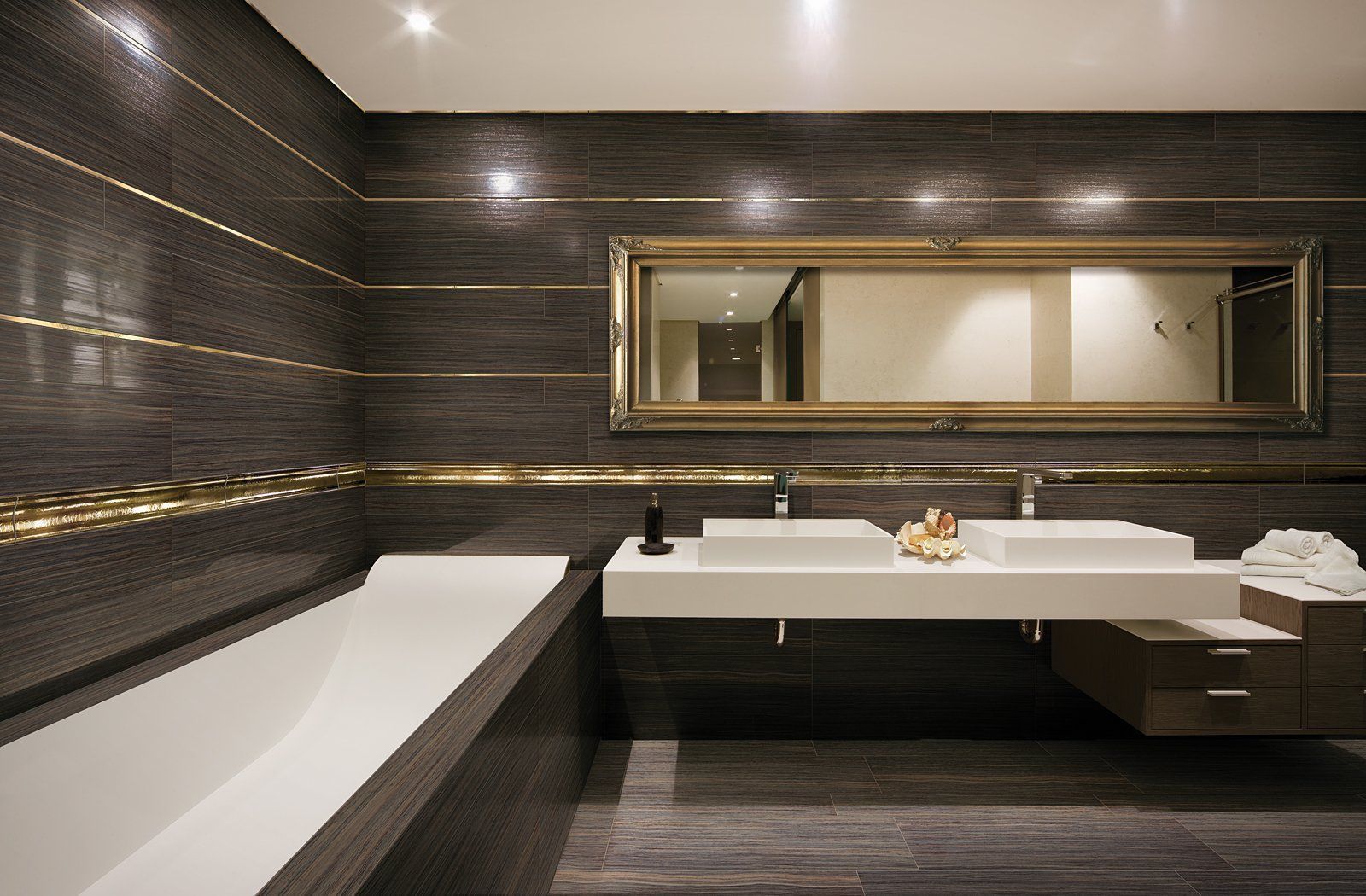 Modern bathroom with dark tile walls, a long mirror, double sink vanity, and a built-in bathtub