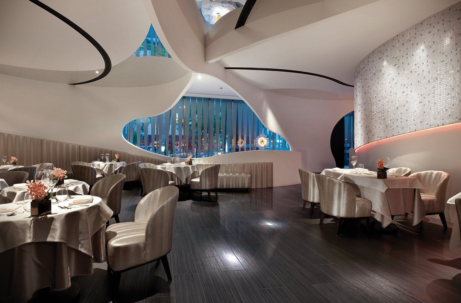 Modern restaurant interior with white sculptural ceiling, dark floor, and set dining tables by blue-lit windows